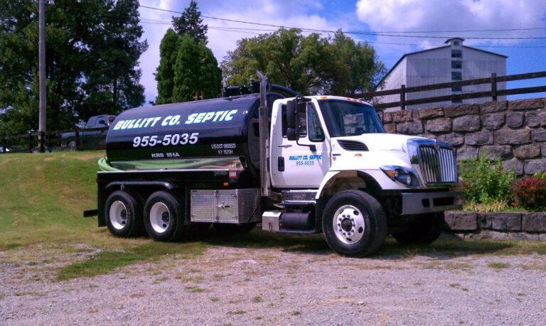 A large septic tank truck parked on a gravel area, with a black tank labeled Bullitt Co. Septic in white lettering. In the background, there are trees and a building. The truck has two axles and tools visible on its side.