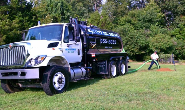 A service truck with a large tank, labeled Bullitt Co. Septic, is parked on a grassy area, while a worker is using a hose to pump waste at a site surrounded by trees.